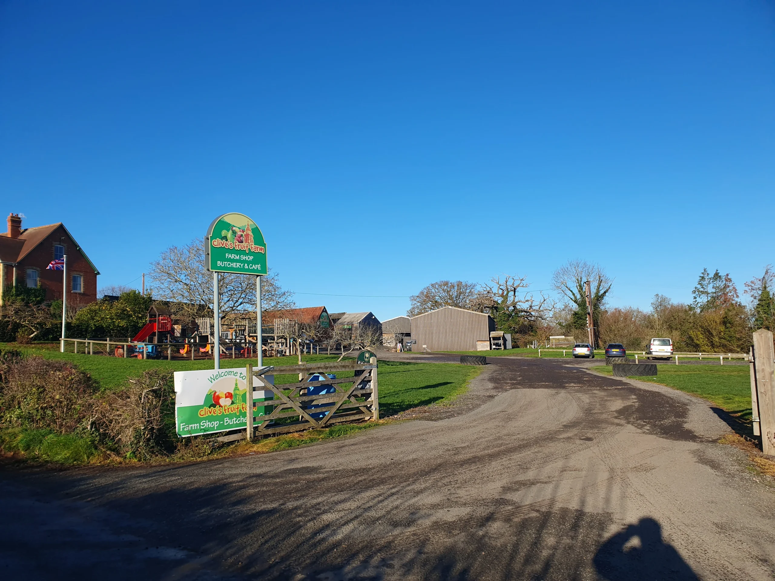 Upton Upon Severn 7 The entrance of Clives Fruit Farm - a tarmac entrance with big open gates, a child play ground and british flag on the left of the field and farm building in the distance.
