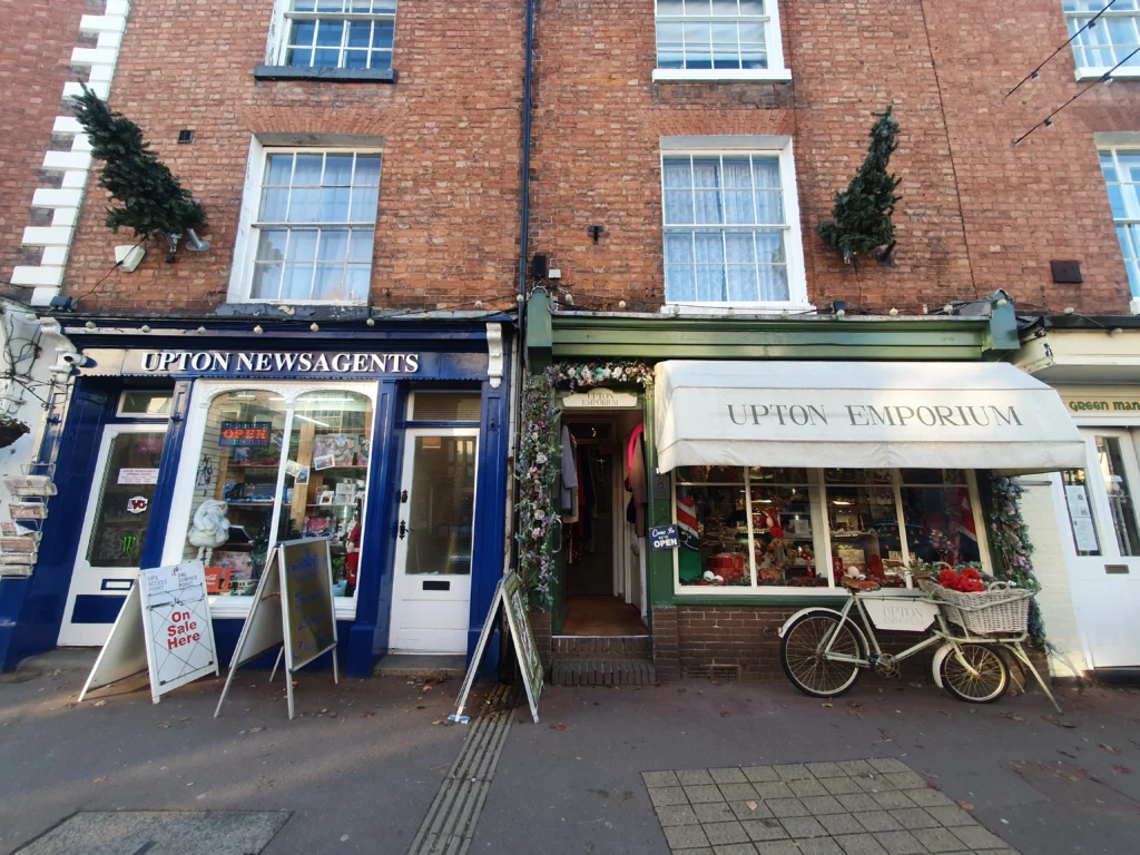 Upton Upon Severn 6 Newagents to the left in blue colour and and gift shop to the right in green - a Christmas trees pointing horizontal and other decorations, the buildings look classical and natural.
